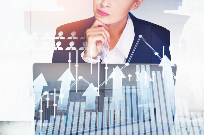 Businesswoman wearing formal suit is working on laptop. New York city skyscrapers in the background. Financial graph, candlesticks and bar diagrams in the foreground. Concept of successful trading